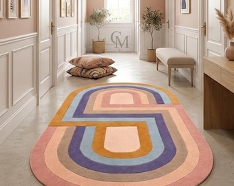 May include: An oval area rug featuring a geometric pattern of concentric rounded shapes in shades of pink, purple, blue, and mustard yellow. The rug is positioned in a hallway with light pink walls, white wainscoting, a wooden console table, and a large arched mirror.