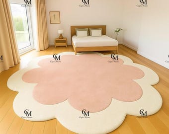 May include: A large, flower-shaped area rug featuring a soft pink center and a cream-colored border. The rug has a plush, textured surface and is positioned on a light-toned wooden floor in a bedroom setting with a bed and curtains visible.