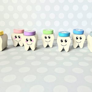 Könnte beinhalten: Eine Sammlung von sieben Holzfiguren in Form von Zähnen mit lächelnden Gesichtern und bunten Kappen. Jeder Zahn ist weiß bemalt mit schwarzen Augen und einem lächelnden Mund. Die Kappen sind in verschiedenen Farben gehalten, darunter Gelb, Rosa, Lila, Grün, Blau und Grau.