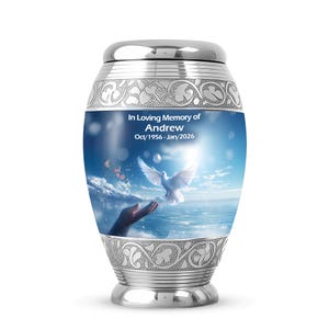 May include: A silver cremation urn with a decorative band and lid. The urn features a serene scene of a dove, hands, and a bright sky with the text "In Loving Memory of Andrew Oct/1956-Jan/2026".
