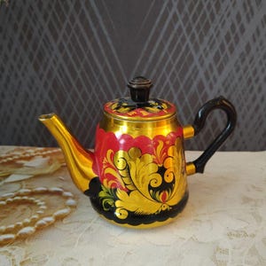 Op de afbeelding: Een decoratieve theepot met een gouden afwerking, met een rood en zwart bloemmotief. De theepot heeft een zwart handvat en deksel en staat op een wit kanten tafelkleed.