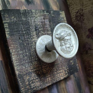 Può includere: Un gancio da parete con una base quadrata in legno tinto scuro e finitura anticata. Al centro è fissato un pomello metallico color argento, decorato con un ritratto in rilievo dettagliato di una figura barbuta vista di profilo.