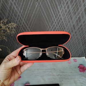 May include: Gold-framed spectacles with a rectangular shape are presented inside an open, bright orange glasses case. The case has a black interior. A black cleaning cloth is visible below the case.