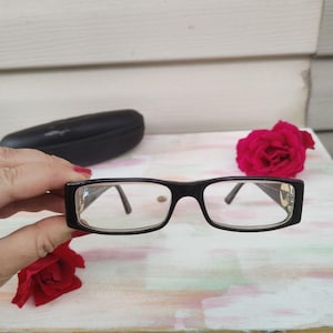 May include: Rectangular eyeglasses with a glossy black frame and clear lenses. The temples feature a small metallic logo emblem near the hinges. The glasses are held in a horizontal position, showcasing the front view of the frame structure.