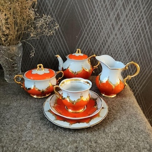 Può includere: Servizio da tè vintage con un design bianco e arancione, rifinito con bordi dorati. Il set include una teiera, zuccheriera, lattiera, tazza e piattino. Il servizio da tè è esposto su una superficie grigia.