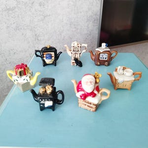 Puede incluir: Una colección de teteras decorativas en miniatura con varios diseños. Las teteras son de diferentes colores, incluyendo negro, marrón, amarillo y blanco. Una tetera presenta un diseño de Papá Noel, y otra tiene una esfera de reloj.