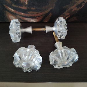May include: Three clear glass door knobs with a faceted, star-like design. Each knob features a silver-toned metal base and a central metal spindle. The glass is transparent and reflects light, showing a multi-pointed geometric pattern.