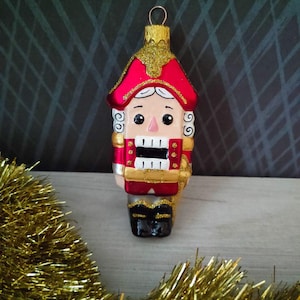 May include: A Christmas ornament shaped like a nutcracker soldier. The ornament has a red hat and jacket with gold accents, a white face, and black boots. The background is a dark gray with a gold garland in the foreground.