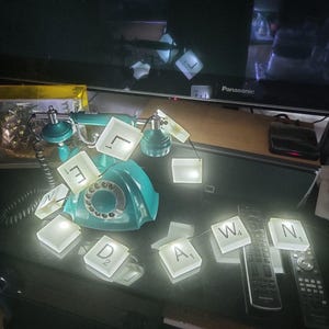 May include: A teal rotary telephone decorated with a string of illuminated white square tiles, each featuring a black letter and a numerical value. The tiles are arranged around the phone, displaying the letters L, E, N, D, A, W, and N.