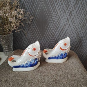 Pode incluir: Duas peças decorativas de cerâmica em forma de peixe. Os peixes são brancos com detalhes laranja nas barbatanas e olhos, e ondas azuis na base. Sobre uma superfície castanha.