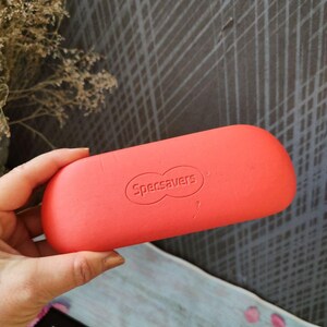 May include: A bright coral glasses case, branded with "Specsavers", held in a hand. The case is oval and has a smooth finish. The background includes a textured grey surface and some dried flowers.