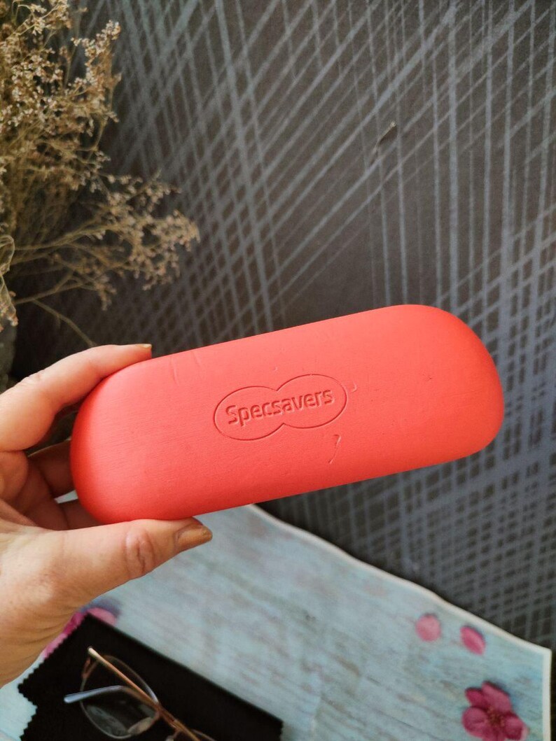 May include: A bright coral glasses case, branded with "Specsavers", held in a hand. The case is oval and has a smooth finish. The background includes a textured grey surface and some dried flowers.