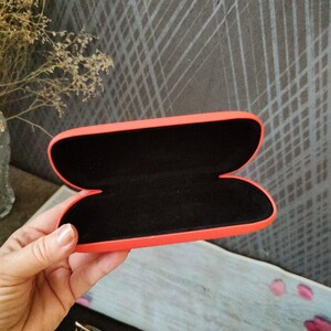 May include: An open, orange glasses case with a black interior is held in a hand. The case is empty, revealing its soft, dark lining. A pair of glasses and a black cleaning cloth are visible below the case.