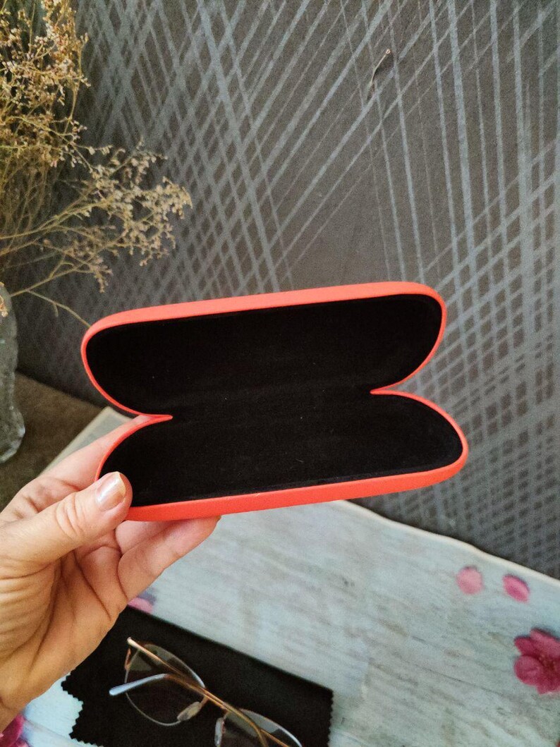 May include: An open, orange glasses case with a black interior is held in a hand. The case is empty, revealing its soft, dark lining. A pair of glasses and a black cleaning cloth are visible below the case.