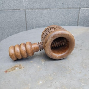 以下が含まれることがあります： 茶色の木製くるみ割り器。円筒形の本体にネジ式の機構と、3つの丸い隆起があるハンドルが付いています。円形の本体の外側には、周囲に複雑な花の彫刻模様が施されています。