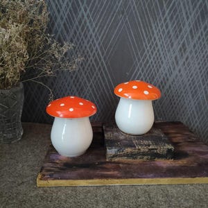 May include: Two ceramic mushroom figurines. The mushroom caps are bright orange with white polka dots, and the stems are white. They are displayed on a wooden surface against a dark gray patterned background. Decorative home decor.