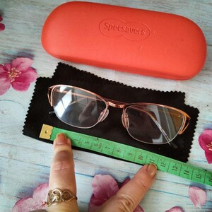 May include: Rose-gold framed eyeglasses on a black cloth, with an orange "Specsavers" glasses case behind them. A green measuring tape is next to the glasses. Pink flowers are scattered around the objects. The glasses have a rectangular shape.