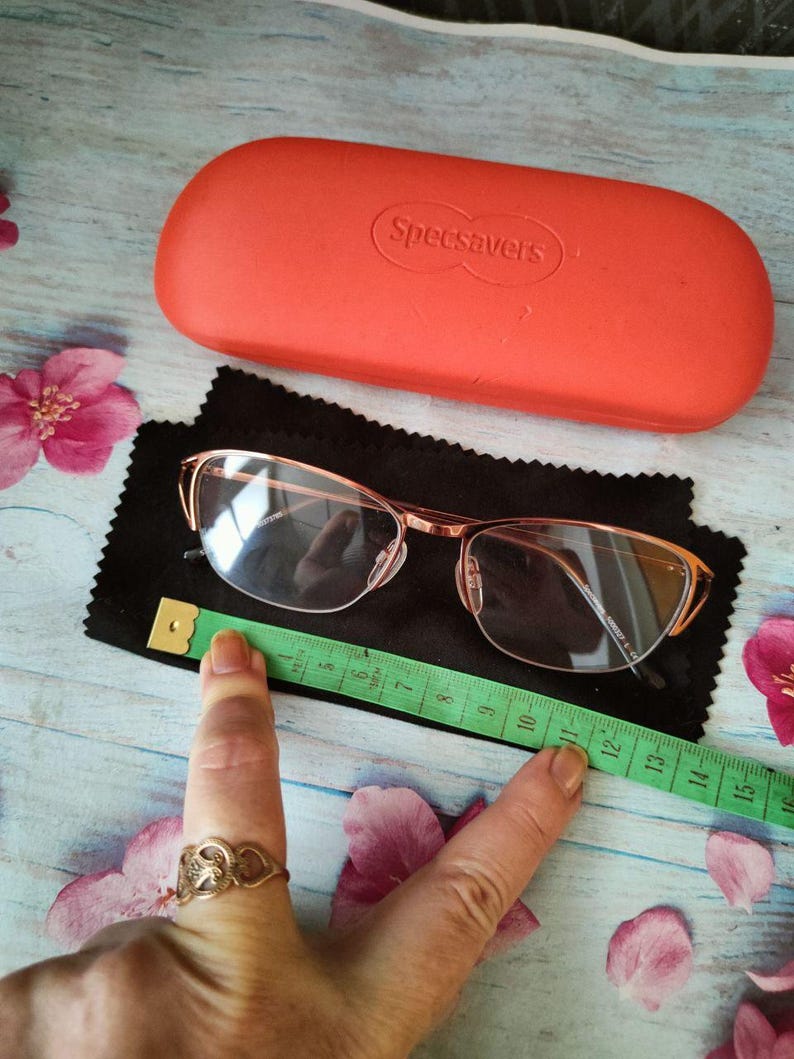 May include: Rose-gold framed eyeglasses on a black cloth, with an orange "Specsavers" glasses case behind them. A green measuring tape is next to the glasses. Pink flowers are scattered around the objects. The glasses have a rectangular shape.