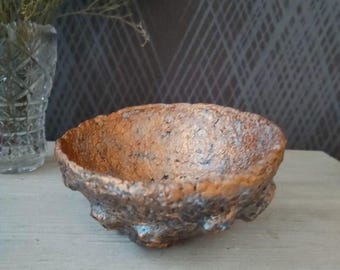 Puede incluir: Un pequeño cuenco de color cobre con una superficie rugosa y texturizada. El cuenco está hecho de un material que se asemeja al papel maché.
