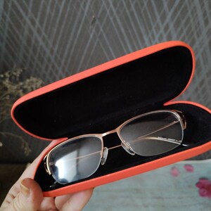 May include: Gold-framed eyeglasses sit in an open, bright orange case with a black interior. The glasses have a rectangular lens shape. The case is being held.