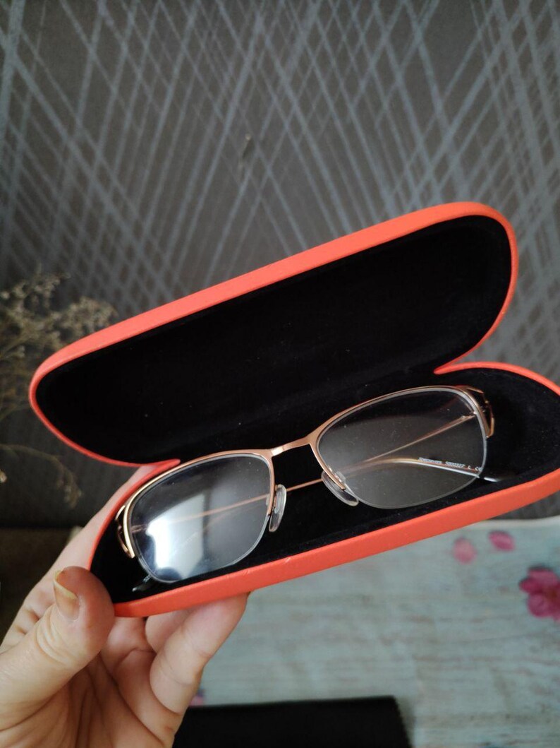 May include: Gold-framed eyeglasses sit in an open, bright orange case with a black interior. The glasses have a rectangular lens shape. The case is being held.