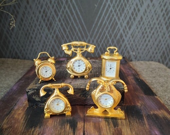 May include: A collection of five miniature gold-toned clocks featuring various designs, including two telephone-shaped clocks, a classic alarm clock, a rectangular carriage clock, and a fish-shaped clock, all with white faces and black hands.