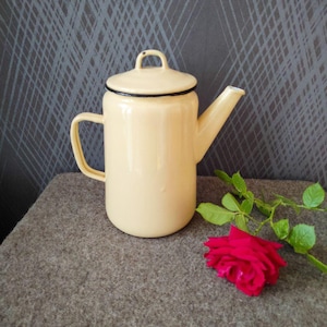 Könnte beinhalten: Eine cremefarbene Emaille-Teekanne mit schwarzem Rand und Griff. Die Teekanne hat einen Deckel mit einem kleinen Schlaufengriff. Eine rote Rose mit grünen Blättern liegt neben der Teekanne auf einer grauen Oberfläche.