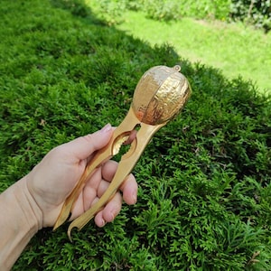 Puede incluir: Un cascanueces de metal dorado con un diseño texturizado similar a una cáscara en la parte superior y dos mangos largos y lisos. La herramienta cuenta con un mecanismo de bisagra con bordes interiores dentados y un acabado en tono dorado pulido.