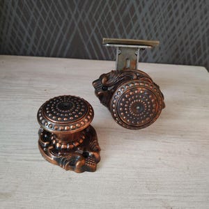 May include: Two antique bronze door knobs with intricate detailing. One knob is upright, the other attached to a metal plate. The knobs feature a circular design with small, raised dots and decorative bases.