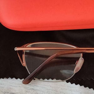 May include: Rose gold eyeglasses with brown temples and clear lenses. The glasses are set against a black cloth and a red glasses case. The glasses have a modern, minimalist design.