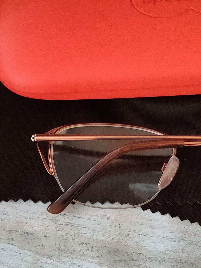 May include: Rose gold eyeglasses with brown temples and clear lenses. The glasses are set against a black cloth and a red glasses case. The glasses have a modern, minimalist design.