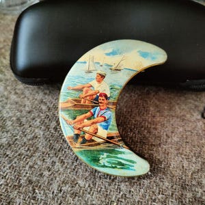 May include: A crescent-shaped brooch featuring a colorful illustration of two individuals rowing wooden boats on water. The scene includes sailboats in the background under a blue sky. The brooch has a metallic gold-toned edge and a smooth surface.