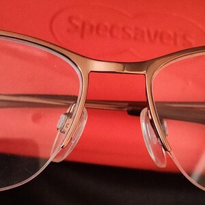 May include: Close-up of gold-framed eyeglasses with clear lenses and nose pads. The glasses are set against a red background. The brand name "Specsavers" is visible in the background.