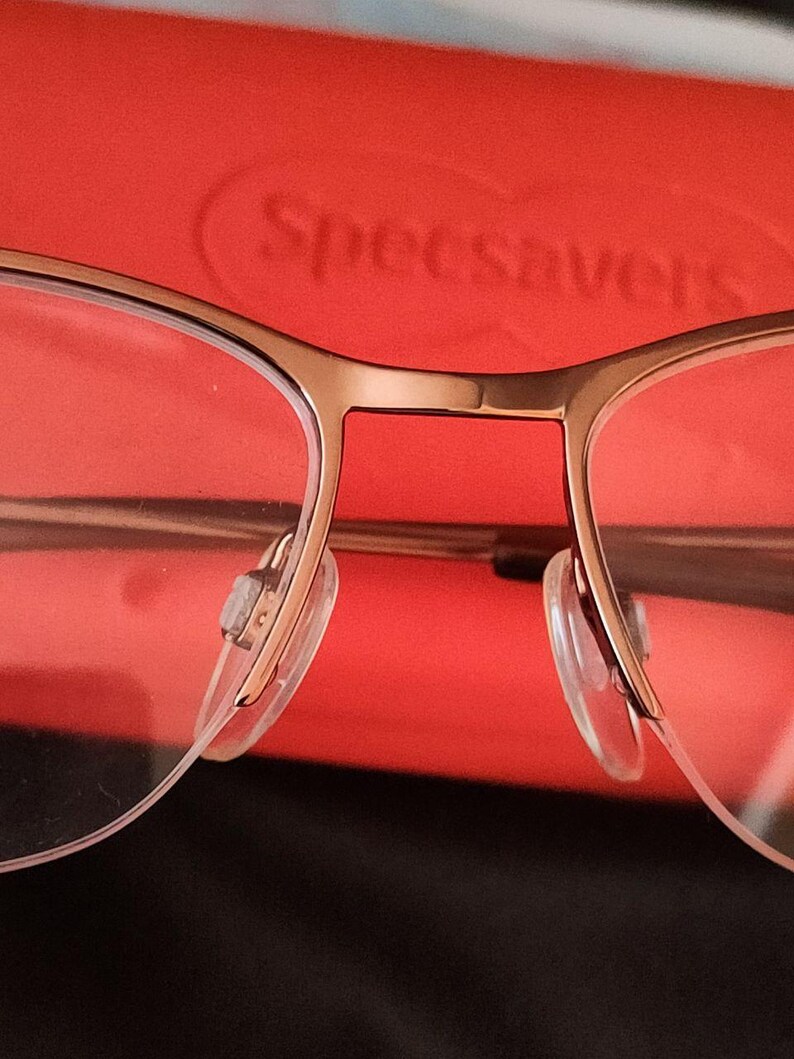 May include: Close-up of gold-framed eyeglasses with clear lenses and nose pads. The glasses are set against a red background. The brand name "Specsavers" is visible in the background.