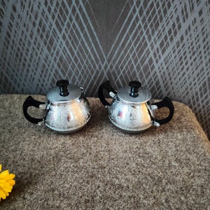 May include: Two silver-colored teapots with black handles and lids. The teapots have an ornate, engraved floral pattern. The teapots are sitting on a brown surface. The background is a dark gray wall with a diamond pattern.