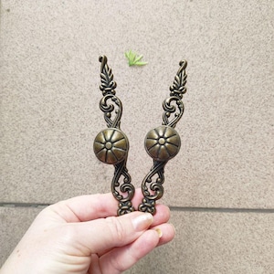 Puede incluir: Dos tiradores de armario de bronce antiguo con diseño floral. Cada tirador presenta un centro redondo en relieve y detalles ornamentados y ondulados. Los tiradores se sostienen en una mano sobre un fondo neutro.