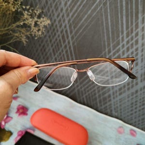 May include: Eyeglasses with a gold-coloured metal frame and clear lenses. An orange glasses case and a black cleaning cloth are visible below. The glasses are held in a hand, against a textured grey background.