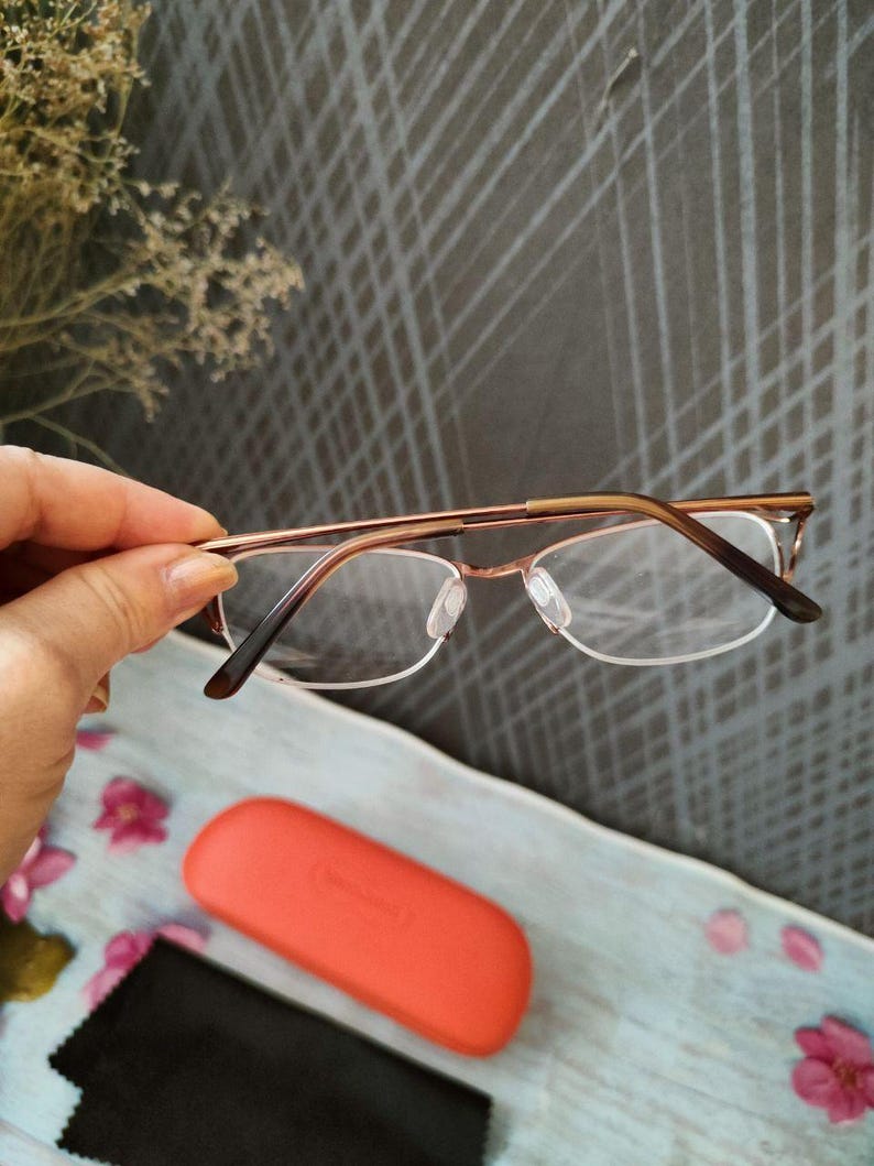 May include: Eyeglasses with a gold-coloured metal frame and clear lenses. An orange glasses case and a black cleaning cloth are visible below. The glasses are held in a hand, against a textured grey background.