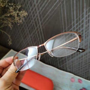 May include: Rose gold eyeglasses with clear lenses are held. The frames have a delicate, modern design. An orange glasses case and a black case are in the background. The background has a gray, textured pattern.