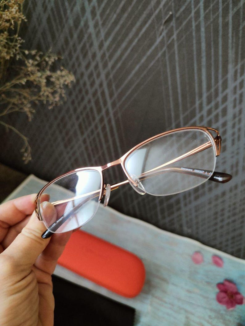 May include: Rose gold eyeglasses with clear lenses are held. The frames have a delicate, modern design. An orange glasses case and a black case are in the background. The background has a gray, textured pattern.
