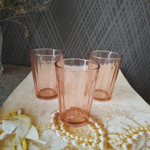 Puede incluir: Tres vasos vintage de color rosa con un diseño acanalado. Los vasos están dispuestos sobre una superficie de encaje color crema, con un collar de perlas y elementos florales decorativos. El fondo presenta un patrón de diamantes grises.