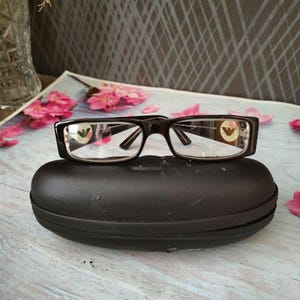 May include: Black rectangular eyeglasses with a dark brown case. The glasses have a black frame and clear lenses. The case is oval and has a matte finish. The glasses are resting on top of the case.