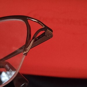 May include: Close-up of eyeglasses with a brown metal frame and clear lenses. The glasses have a unique design with a curved top and a sleek, modern aesthetic. The background is a solid red colour.