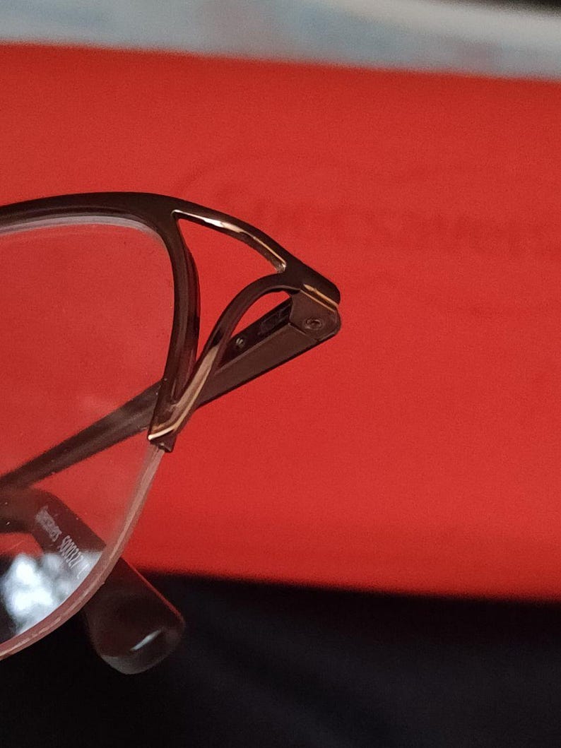 May include: Close-up of eyeglasses with a brown metal frame and clear lenses. The glasses have a unique design with a curved top and a sleek, modern aesthetic. The background is a solid red colour.