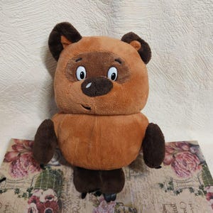 Peut inclure: Une peluche en forme d'ours brun, avec des oreilles, des pattes et des traits du visage brun foncé. L'ours a une expression amicale avec de grands yeux et un petit sourire. Il est posé sur une surface à motifs floraux.