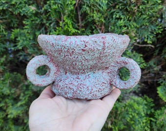 Puede incluir: Un pequeño cuenco de cerámica texturizada con un acabado moteado rojo y azul. El cuenco tiene dos asas circulares y se sostiene en una mano sobre un fondo de follaje verde. El cuenco está diseñado para fines decorativos.
