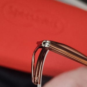 May include: Close-up of spectacles with a brown frame and silver hinges. The glasses have a curved design and are set against a red background. The focus is on the details of the frame and the hinge.