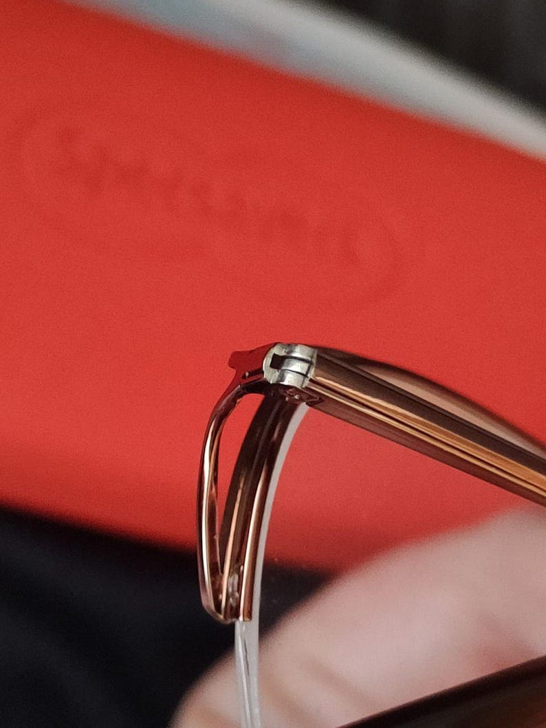 May include: Close-up of spectacles with a brown frame and silver hinges. The glasses have a curved design and are set against a red background. The focus is on the details of the frame and the hinge.
