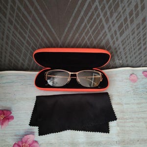 May include: Gold-rimmed spectacles in an open orange case with a black interior. A black microfiber cleaning cloth is beside the case. The background is a light blue surface with pink flowers and a dark grey patterned backdrop.