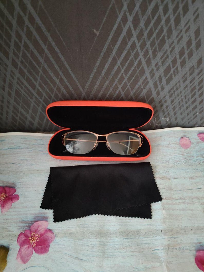 May include: Gold-rimmed spectacles in an open orange case with a black interior. A black microfiber cleaning cloth is beside the case. The background is a light blue surface with pink flowers and a dark grey patterned backdrop.
